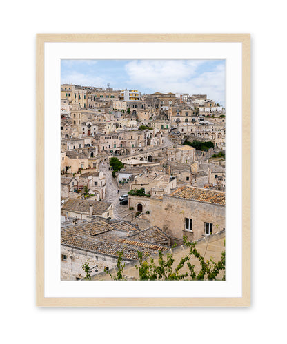 Vertical architecture photograph of Matera, Italy featuring textured stone buildings and neutral tones in a light wood frame.