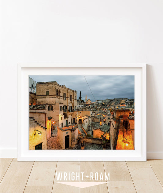 Horizontal twilight photograph of Matera, Italy with glowing amber streetlights and historic stone buildings in white frame against a white background