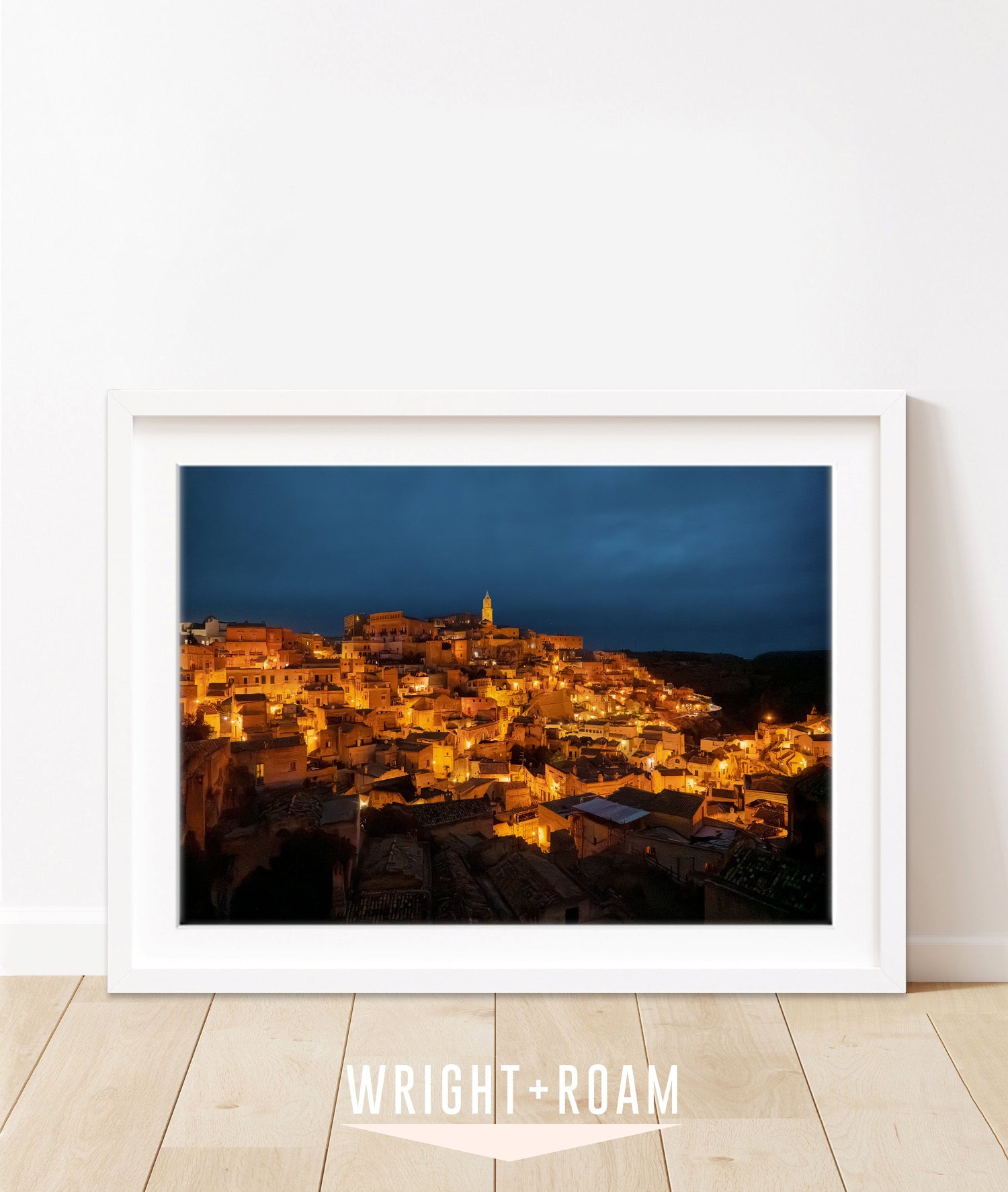 framed horizontal night photograph of Matera, Italy with glowing amber city lights against a navy sky.