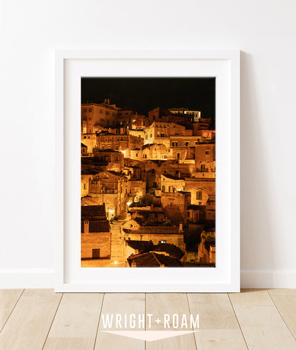 framed photograph of Historic city of Matera, Italy at night with glowing amber lights and dark stone architecture in a vertical cityscape photograph.