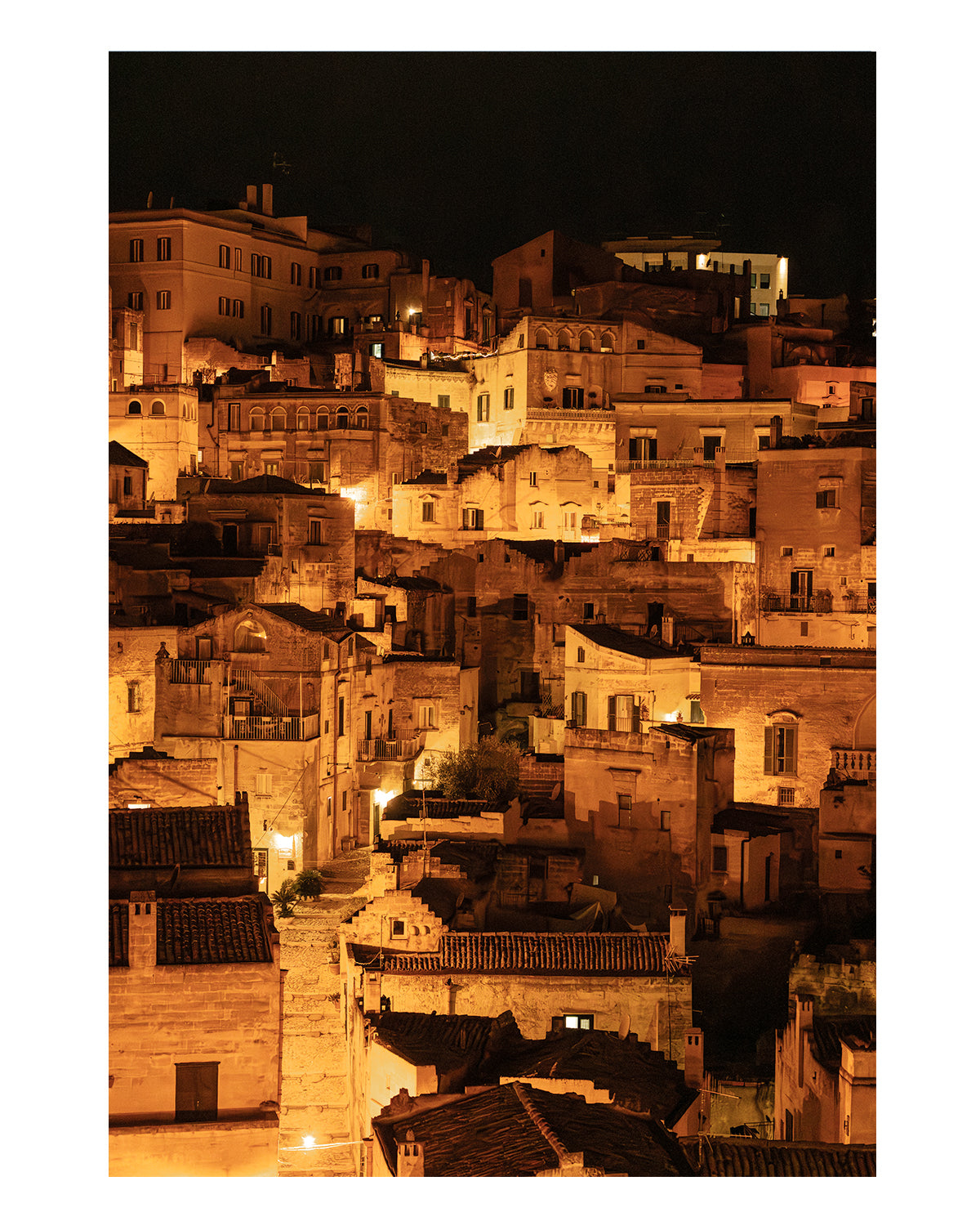 unframed photograph of Historic city of Matera, Italy at night with glowing amber lights and dark stone architecture in a vertical cityscape photograph.