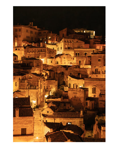 unframed photograph of Historic city of Matera, Italy at night with glowing amber lights and dark stone architecture in a vertical cityscape photograph.
