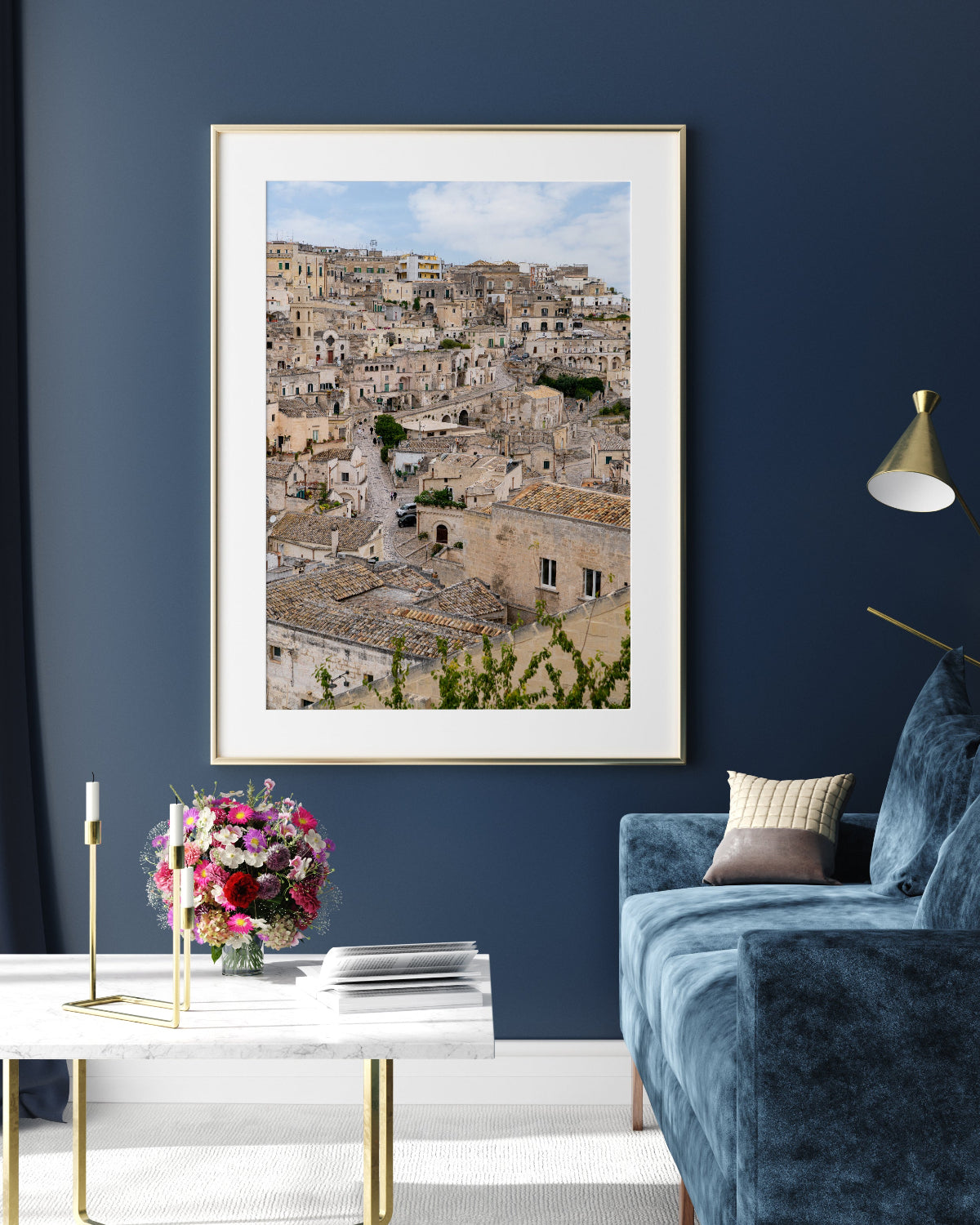 Framed artwork of a cityscape on a blue wall above a white table with flowers and books, next to a blue sofa.