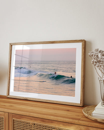 framed wall art featured soft pink sunrise over the ocean with surfers surfing. Framed art on Wood dresser.