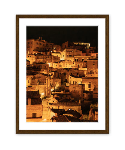 dark wood frame fine art photograph of Historic city of Matera, Italy at night with glowing amber lights and dark stone architecture in a vertical cityscape photograph.