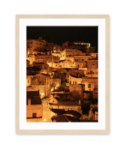 light wood frame photograph of Matera, Italy at night with glowing amber lights and dark stone architecture 